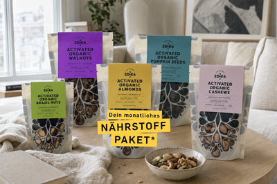 Fünf Beutel aktivierte Bio-Nüsse von 2DiE4 auf einem Couchtisch im Wohnzimmer, davor eine Schale mit Nüssen, Banner Dein monatliches Nährstoff Paket