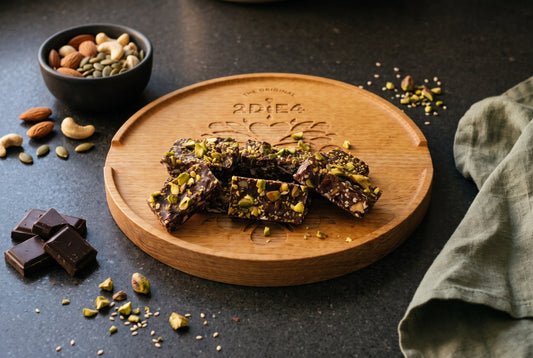Nut Crunch Bites mit gehackten Pistazien auf einem runden 2DiE4-Eichenbrett, daneben eine Schale mit Mandeln, Cashews und Kürbiskernen und Stücke Zartbitterschokolade